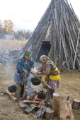 Two Khanty women cooking outside in the autumn at a camp on the Synya River. Behind them is a wooden store. Yamal, Western Siberia, Russia